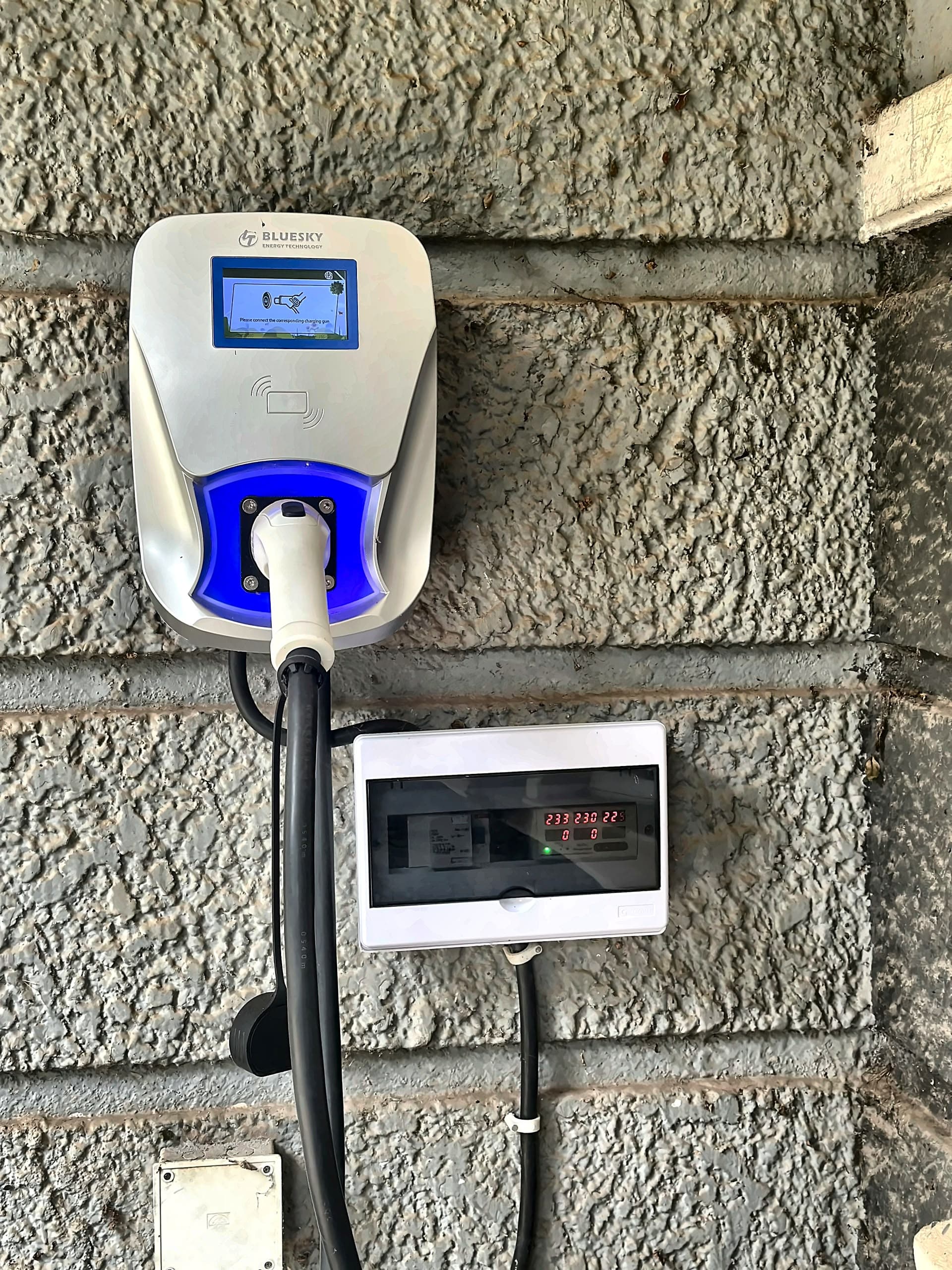 A wall-mounted EV charger with a touchscreen, RFID pad, and glowing blue status light, plugged in beside its electrical panel